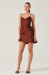 A woman is wearing a silky, satin-like two-piece outfit in a deep reddish-brown color. The top is a camisole with thin spaghetti straps and a v-neckline edged with black lace trim. The matching skirt is short, with a slight A-line shape and a subtle asymmetrical hem. She is also wearing black open-toe high heels.